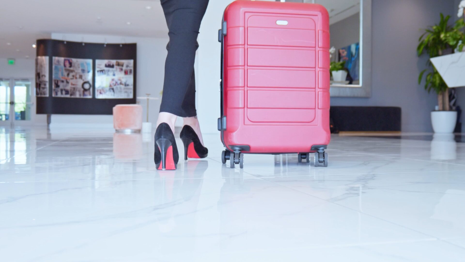 Frame from the Karol Hotel film: a traveler in black heels rolling a red suitcase across the lobby's marble floor.